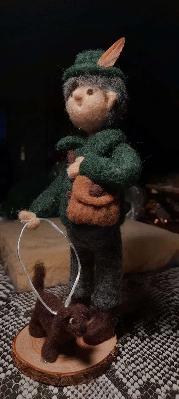 Filzfigur mit grünem Federhut, Umhängetasche und kleinem Hund auf einer Holzscheibe.