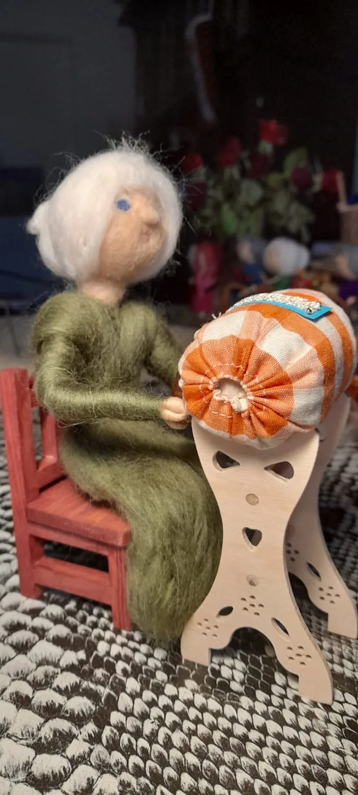 Sitzende Filzfigur im grünen Kleid vor einem orange-weiß karierten Handarbeitskissen auf einem Holzständer.