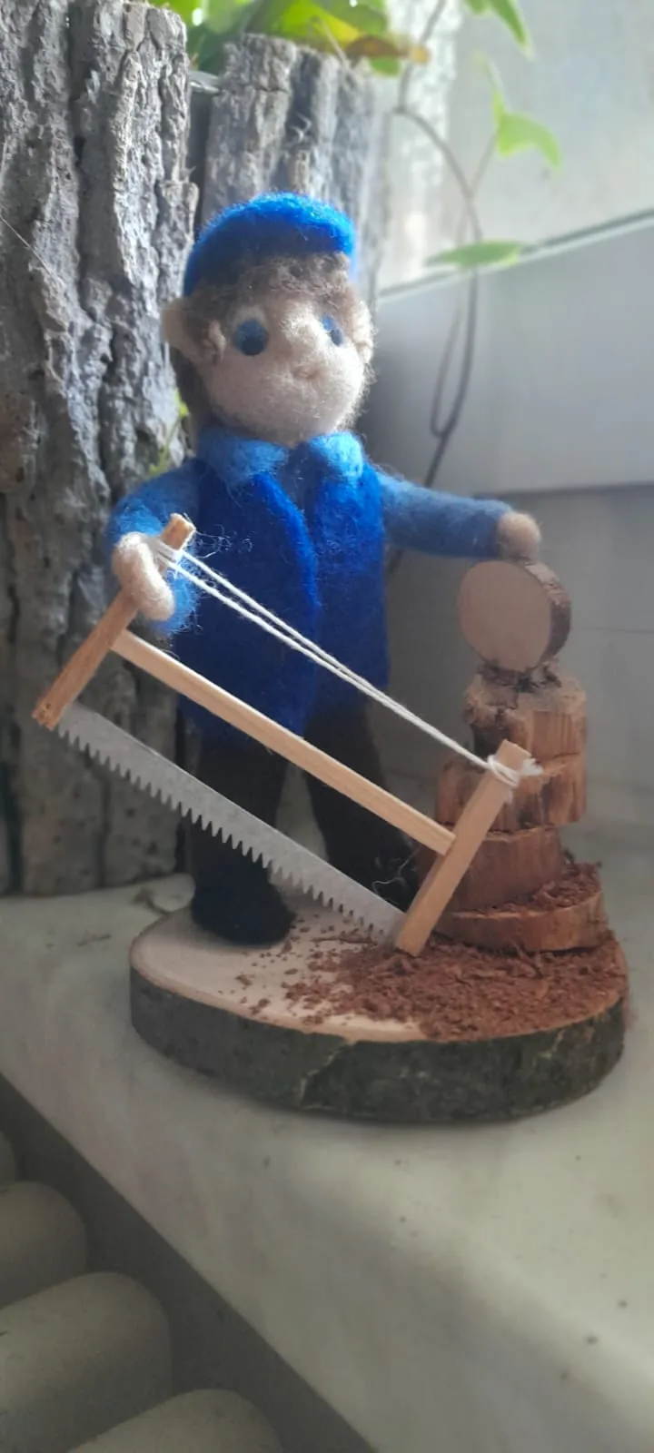 Blau gekleidete Filzfigur mit einer Gestellsäge neben einem Holzblock auf einer Holzscheibe.