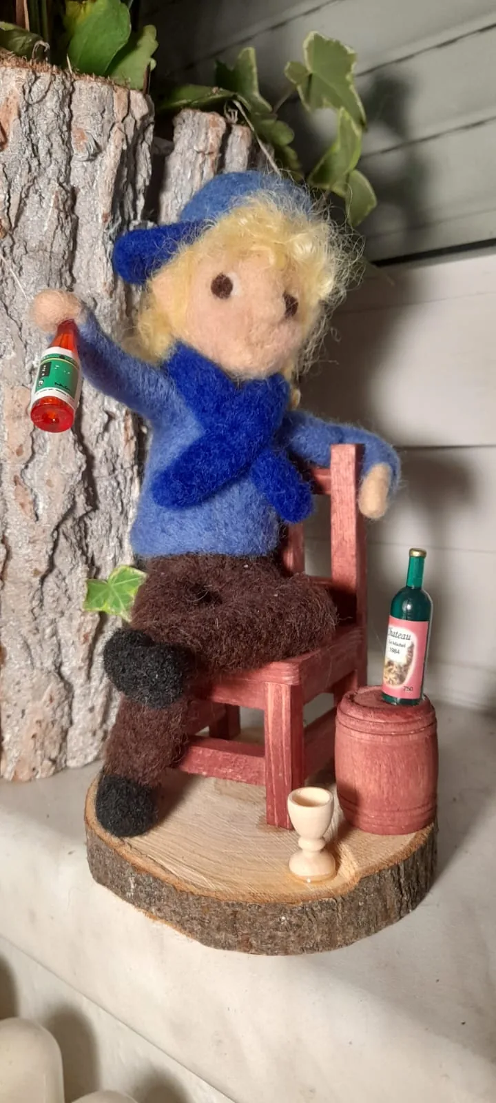 Sitzende Filzfigur mit blauer Mütze, kleinen Flaschen und einem Becher neben einem Fass.